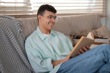 Un chico joven con gafas sentado en un sofa de la casa de una persona leyendole un libro